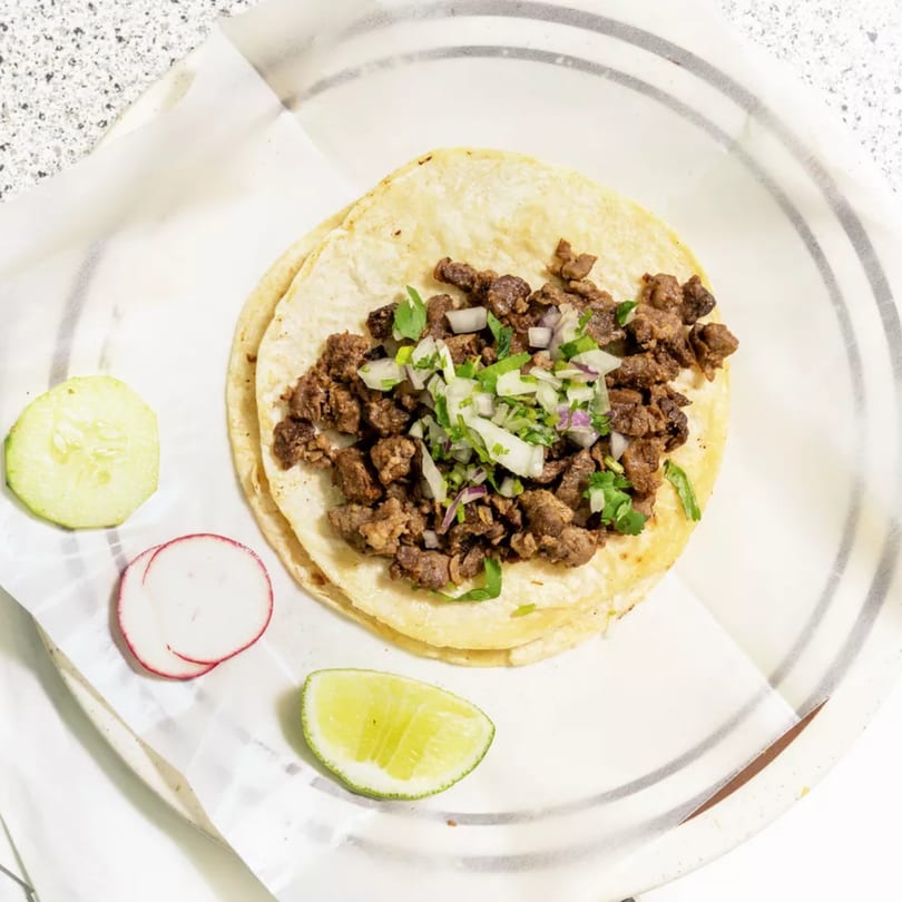 Two street tacos with chopped meat, onions, and cilantro, served with lime, cucumber, and radish.