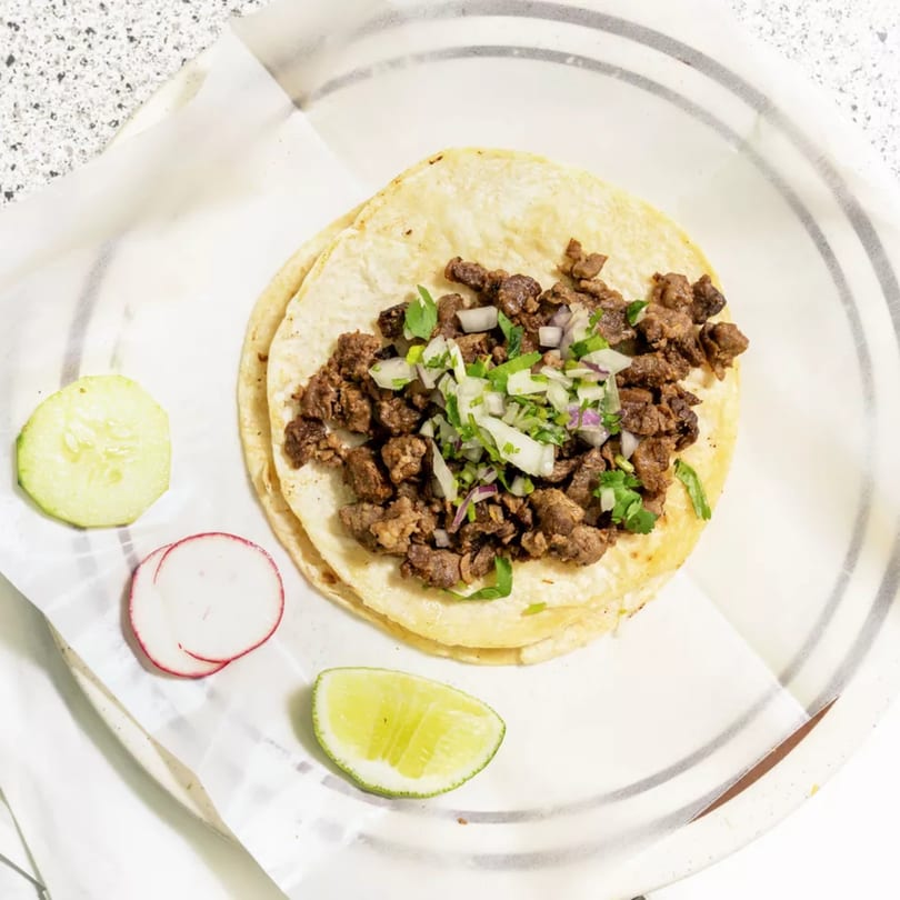 Two Mexican street tacos with carne asada, cilantro, and onions, served with lime, cucumber, and radish.