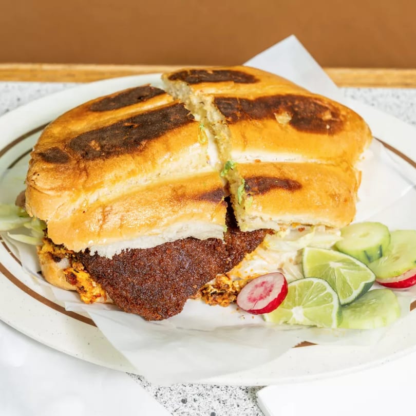 A large, toasted Mexican torta sandwich, cut in half, with a side of lime, cucumber, and radish.