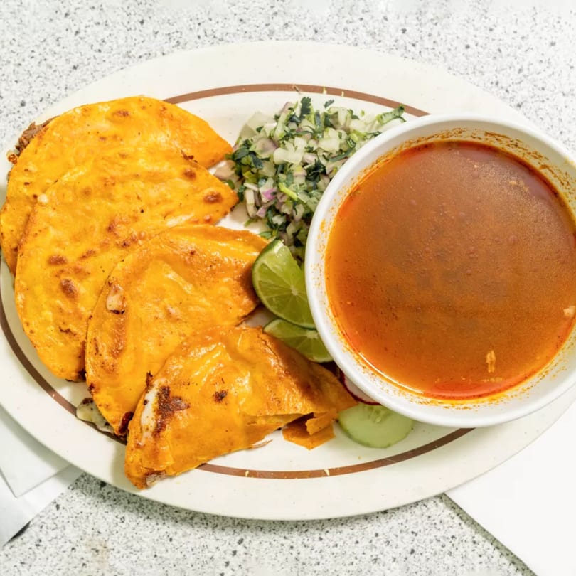 A plate of four golden-brown birria tacos with a side of chopped onions, cilantro, lime, and a bowl of consommé.