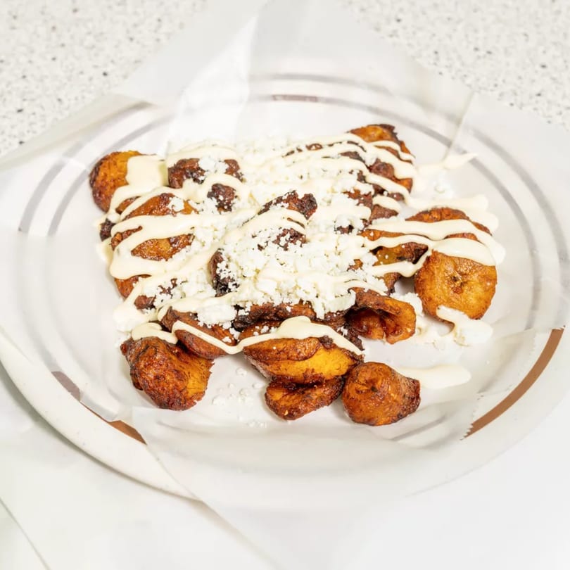 Fried plantains drizzled with white sauce and sprinkled with crumbled white cheese on a white plate.