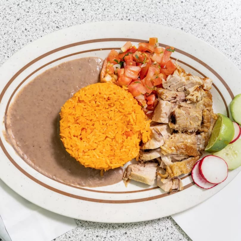 A plate of carnitas with rice, refried beans, pico de gallo, lime, cucumber, and radish.