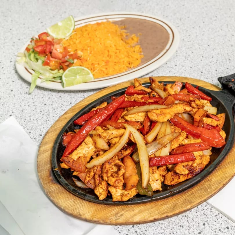 Sizzling chicken fajitas with red peppers and onions, alongside a plate of rice, beans, and salad.