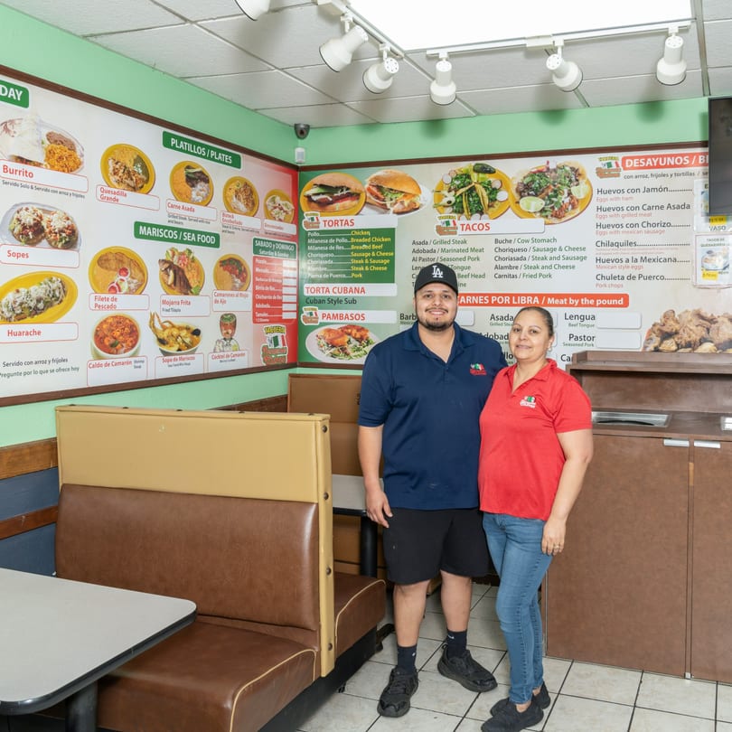 Two restaurant staff members stand proudly in front of a large menu board at Taqueria Hermanos Chavez.