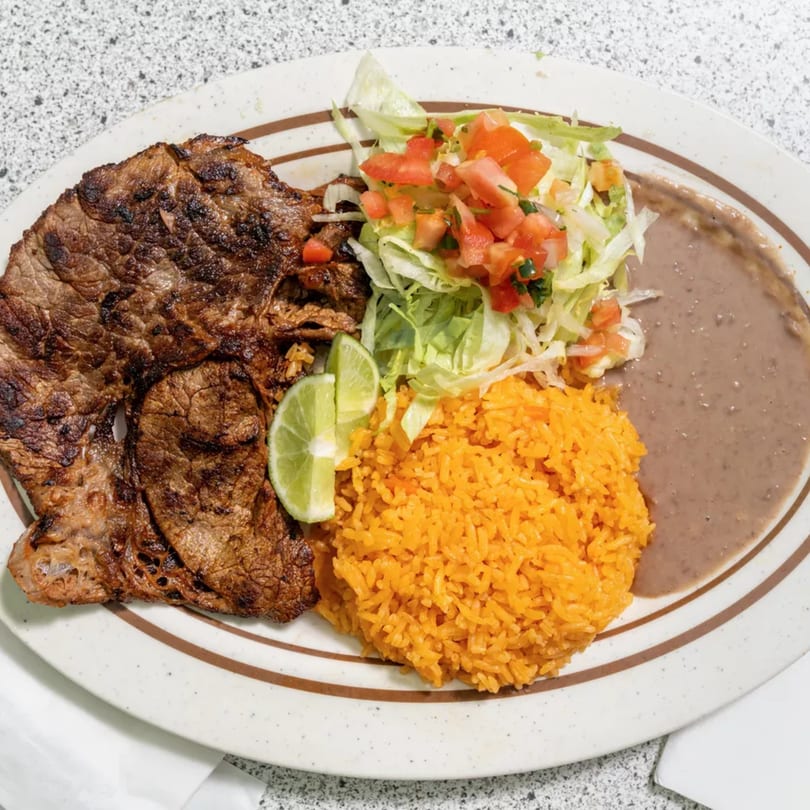 Grilled steak, orange rice, refried beans, and a fresh salad with lime on a white plate.